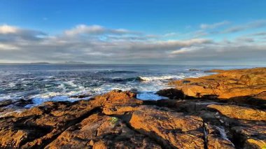 Berrak mavi gökyüzünün altındaki engebeli kayalara çarpan dalgaların çarpıcı görüntüsünü hissedin.