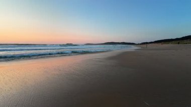 Güzel, sakin bir sahil sahnesi baş döndürücü bir gün batımı ve sakin dalgalar gökyüzündeki yumuşak renkleri yansıtıyor.