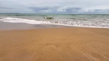 Yumuşak ve ılık kumları, kıyıya vuran hafif dalgaları ve yukarıda bulutlu bir ufku tasvir eden sakin bir sahil sahnesi.