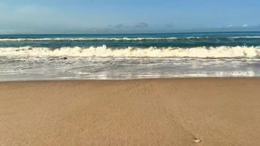 Yumuşak, rahatlatıcı dalgalar yukarıdaki açık mavi gökyüzünün altındaki kumlu kıyıya hafifçe çarpıyor.