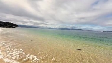 Bu sakin sahil sahnesi, bulutlu ve bulutlu bir gökyüzünün altında yumuşak kumları ve mükemmel sakin suları sergiliyor.