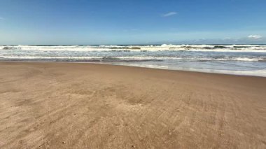 Sakin bir sahil sahnesi, yumuşak dalgalar parlak mavi bir gökyüzü ve yumuşak pamuklu bulutların altında kıyıları sarıyor.