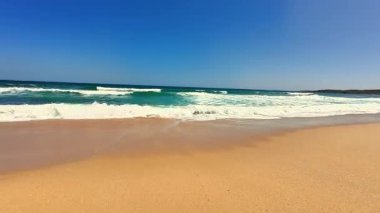 Yumuşak, sıcak kumları, dalgaları ve yukarıda canlı mavi gökyüzünü gösteren nefes kesici bir sahil manzarası.