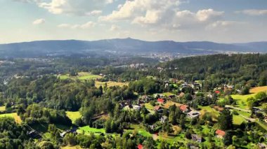 Parlak, güneşli bir gökyüzünün altında tepeler ve büyüleyici evlerle çevrili yeşil bir vadinin çarpıcı manzarası.