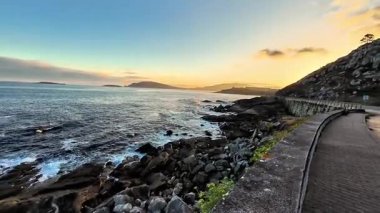 Gün batımında nefes kesen güzel bir sahil yolu, dalgalı okyanus dalgaları ve etrafındaki engebeli uçurumları gösteriyor.