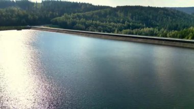 Çerçevelenmiş yemyeşil ve pitoresk tepeler tarafından çerçevelenmiş, altın güneş ışığını yansıtan büyük bir rezervuarın huzurlu manzarası.