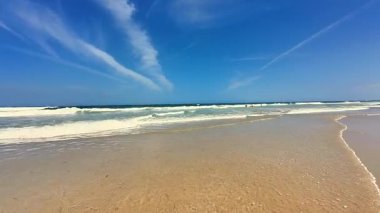 Huzurlu ve sakin bir sahil sahnesi yumuşak altın kum, berrak nazik dalgalar ve parlak mavi bir gökyüzü sergiliyor.