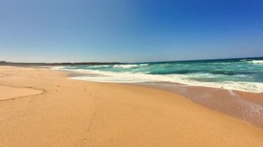 Güzel, sakin bir sahil sahnesi yumuşak altın kum, yumuşak dalgalar ve parlak mavi gökyüzü sergileniyor.