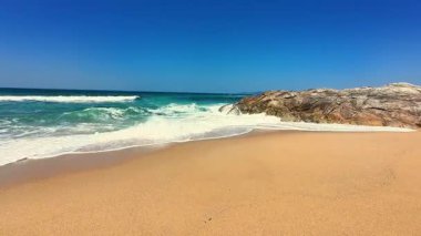 Yumuşak kum, yumuşak dalgalar ve açık bir gökyüzünün altında büyük bir kaya oluşumunun yer aldığı pitoresk bir sahil sahnesi..