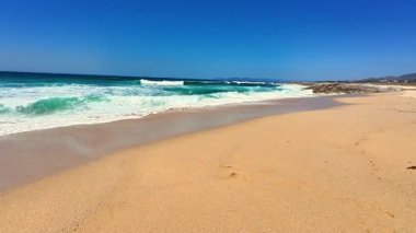 Açık mavi gökyüzünün altında altın kumlar ve canlı turkuaz dalgalar sergilenen nefes kesici bir plaj sahnesi.