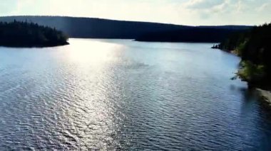 Parlak bir gökyüzünün altında yoğun ve canlı ormanlarla çevrili güzel, sakin bir göl manzarası.
