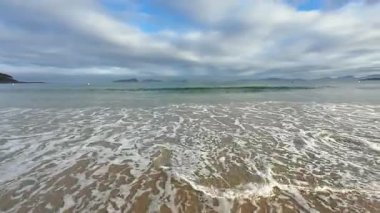 Yumuşak dalgaları ve kasvetli gökyüzünü güzel bir şekilde yansıtan sakin bir sahil manzarası, seyahat ya da doğa teması için ideal.