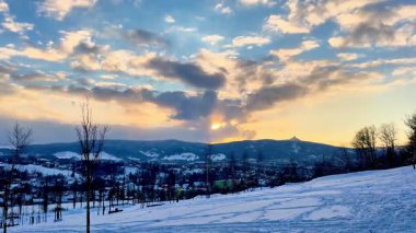 Nefes kesen kış günbatımı dramatik bulutlarla süslenmiş sakin karlı bir manzarayı canlı bir şekilde aydınlatıyor.