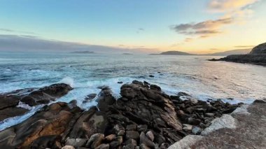 Gün batımında güzel, sakin bir kıyı manzarası, dalgaların ritmik bir şekilde kayalık bir kıyıya çarpması.