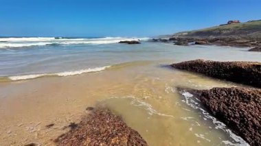 Şaşırtıcı derecede güzel bir plaj sahnesi açık mavi gökyüzünün altında yavaşça dalgalar ve engebeli kayalar sergiliyor.