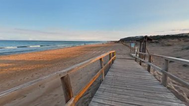 Sakin, ahşap bir patika gün batımında kumlu bir sahile çıkar. Sakin okyanus dalgaları yavaşça kıyıya vurur.