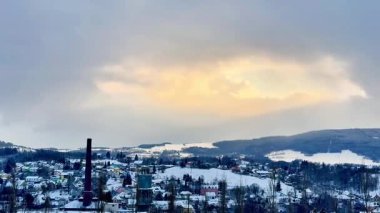 Huzurlu bir kış sahnesi karla kaplı bir kasabayı kasvetli bulutların altında uzak bir baca yığınıyla gösteriyor.