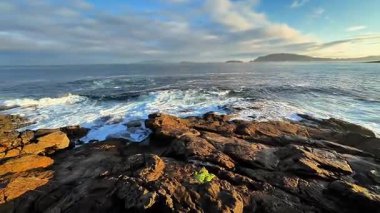 Sakin bir sahil sahnesi, berrak mavi gökyüzünün altında engebeli kayalara çarpan büyüleyici dalgaları gösteriyor.