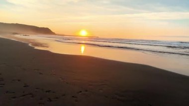Gün batımında sakin ve sakin bir sahil sahnesi yumuşak dalgalar sergiliyor ve her yerde huzurlu bir atmosfer yaratıyor.