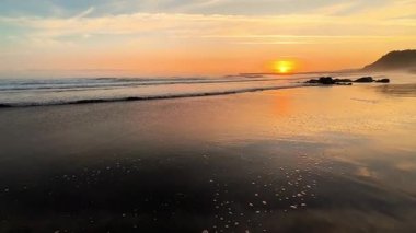 Bu, sakin sahil dalgalarını yansıtan güzel bir gün batımının büyüleyici bir görüntüsü.