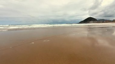 Huzurlu bir sahil sahnesi bulutlu bir gökyüzünün altında yumuşak dalgaların kumlu sahile hafifçe vuruşunu tasvir ediyor.