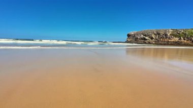 Güzel ve sakin bir sahil sahnesi altın kum, nazik dalgalar ve geniş mavi bir gökyüzü sergiliyor.