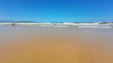 Yumuşak, altın kum, yumuşak dalgalar ve berrak mavi gökyüzü içeren sakin ve sakin bir sahil sahnesi.