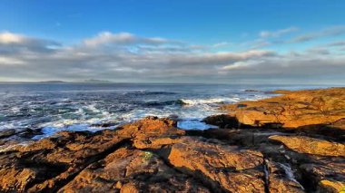 Berrak mavi gökyüzünün altındaki engebeli kayalara çarpan dalgaların çarpıcı görüntüsünü hissedin.