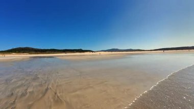 Huzurlu bir sahil sahnesi, yukarısında açık mavi gökyüzü ile kıyıya vuran sakin, dingin dalgalar.