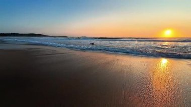 Sakin bir sahil günbatımı, sakin sularda altın yansımalar ve kıyıya vuran yumuşak dalgalar sergiliyor.