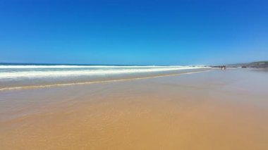 Göz kamaştırıcı bir plaj sahnesi yumuşak altın kum, kıyıya vuran nazik dalgalar ve yukarıda parlak mavi bir gökyüzü.