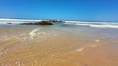 Geniş kumlu sahilleri ve berrak mavi gökyüzünün altında narin okyanus dalgalarını gösteren sakin bir sahil.