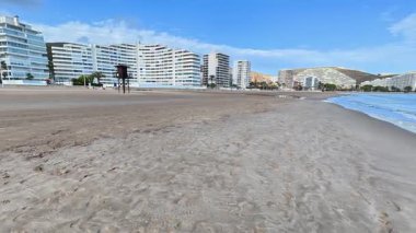 Sakin bir şehir plajı sahnesi canlı, berrak mavi gökyüzüne karşı modern binalar sergiliyor.