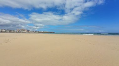 Güzel, sakin bir sahil sahnesi altın kumlar ve parlak mavi bir gökyüzünün altında parıldayan nazik dalgalar sergiliyor.