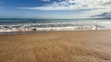 Huzurlu ve sakin bir sahil sahnesi yumuşak, sıcak kum, hafif dalgalar ve yukarıda açık mavi bir gökyüzü barındırır.