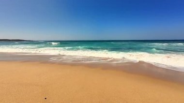 Sakin ve sakin bir sahil sahnesi. Yumuşak dalgaların, açık mavi bir gökyüzünün altında yumuşak altın kuma vurduğu.