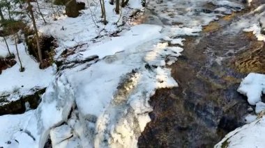 Güzel, sakin bir kış sahnesi, karla kaplı bir manzara ve ağaçlarla sarılmış, nazikçe akan bir dereyi gözler önüne seriyor.