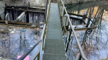 Metal bir köprü, enkazla çevrili sakin suyu gerer, endüstriyel bozulmaları ve çevresel ihmalleri gösterir.