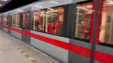 Modern bir metro treni, çeşitli yolcular ve şehir yaşamını ön plana çıkaran kalabalık bir şehir istasyonuna ulaşır.