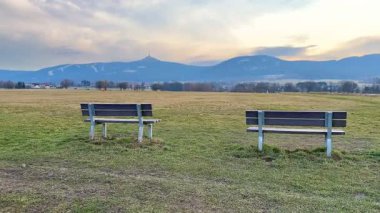 Bu, bulutlu bir gökyüzünün altında uzak dağlara bakan iki bankın yer aldığı huzurlu ve resimsel bir manzara.