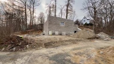 Tamamlanmamış bir bina, ağaçlar ve dağınık inşaat atıklarıyla çevrili verimli bir alanda yer almaktadır.