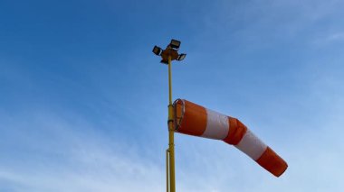 Şaşırtıcı derecede açık mavi bir gökyüzü, uzun bir direğe monte edilmiş çeşitli ışıklandırma ekipmanlarıyla birlikte renkli bir rüzgar çorabı sergiliyor.