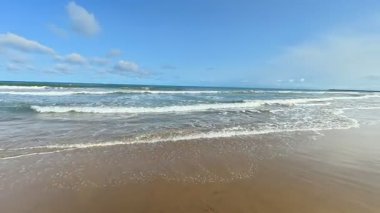 Resimli ve sakin bir sahil sahnesi, parlak mavi gökyüzünün altında süzülen nazik dalgaları sergiliyor.