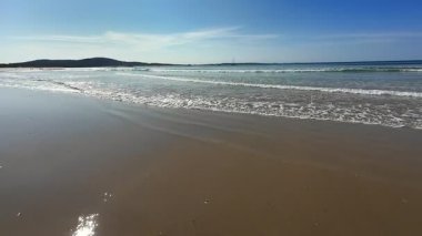 Sakin bir sahil, hafif dalgalar kıyıya vuruyor, parlak mavi bir gökyüzü altında, rahatlamak için mükemmel.