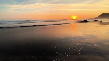 Bu, sakin sahil dalgalarını yansıtan güzel bir gün batımının büyüleyici bir görüntüsü.