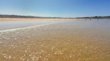 Sakin bir sahil sahnesi, berrak mavi gökyüzü altında yumuşak dalgalar, rahatlamak ve gevşemek için mükemmel.