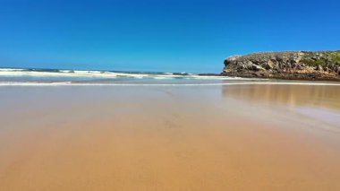 Güzel ve sakin bir sahil sahnesi altın kum, nazik dalgalar ve geniş mavi bir gökyüzü sergiliyor.