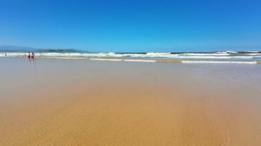 Yumuşak, altın kum, yumuşak dalgalar ve berrak mavi gökyüzü içeren sakin ve sakin bir sahil sahnesi.