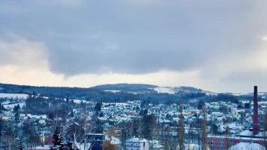 Arka planda yükselen görkemli sisli dağlarla kaplı karla kaplı bir kasabanın sakin kış manzarası.