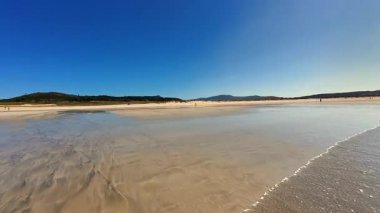 Huzurlu bir sahil sahnesi, yukarısında açık mavi gökyüzü ile kıyıya vuran sakin, dingin dalgalar.